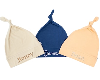 Könnte beinhalten: Drei Baby-Knotenmützen in Creme, Marineblau und Pfirsich. Jede Mütze hat einen aufgestickten Namen auf der Vorderseite: "Jimmy", "James" und "Bean". Die Mützen sind aus weichem Stoff und haben eine geknotete Oberseite.