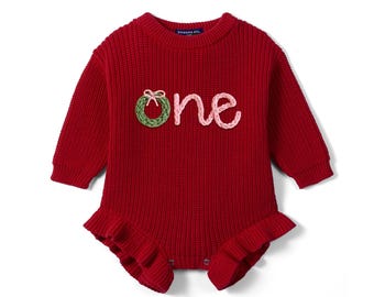 May include: A long-sleeved red knit romper featuring a ribbed texture and a ruffled hem. The front displays the word one in textured yarn, with the letter o replaced by a green wreath with a small pink bow and the letters ne in light pink.