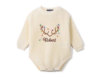 May include: A cream-colored long-sleeve knit romper featuring an embroidered design of brown reindeer antlers draped with colorful string lights. The name Robert is embroidered in brown script below the antlers on the front of the garment.