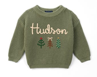 May include: An olive green chunky knit sweater featuring the name Hudson embroidered in cream-colored yarn across the chest. Below the name are three small embroidered Christmas tree designs in green, red, and brown with a cream bow detail.