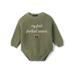 May include: An olive green long-sleeve knit romper featuring a crew neckline and ribbed texture. The front displays the white embroidered text 'my first football season' positioned above a brown and white graphic of an American football.