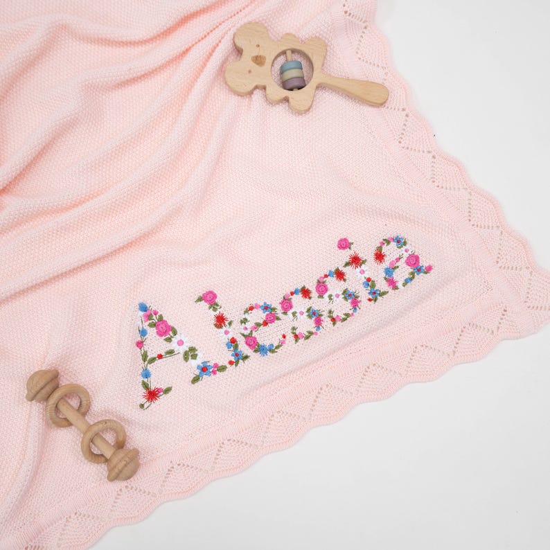 May include: A light pink baby blanket with the name "Aleesia" embroidered in colorful flowers. The blanket has a scalloped edge and is accompanied by two wooden baby toys.