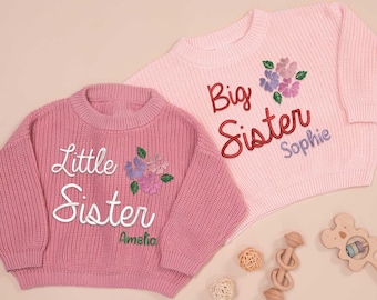 May include: Two pink knit sweaters, one darker than the other. The darker sweater says "Little Sister Amelia" with floral embroidery. The lighter sweater says "Big Sister Sophie" with floral embroidery. Wooden toys and decorative balls are scattered around.