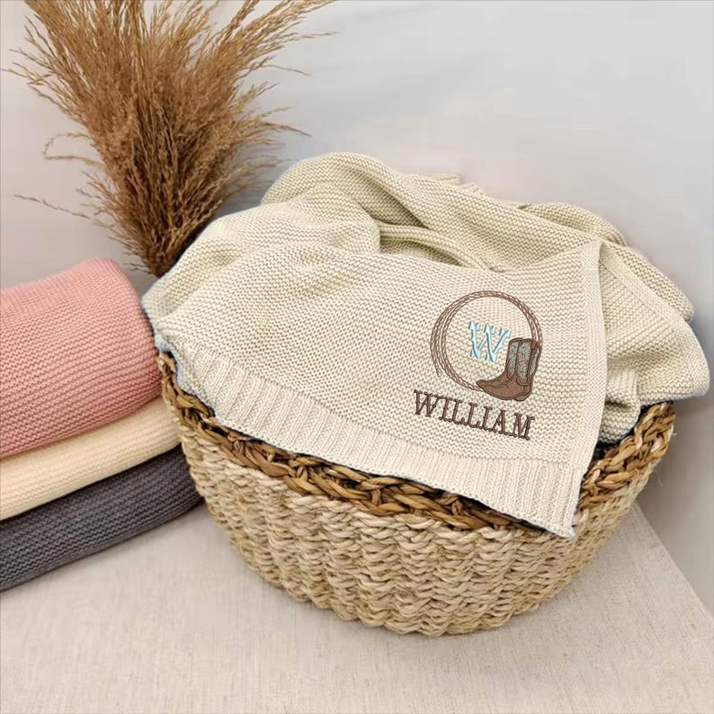 Puede incluir: Una manta beige tejida con una bota de vaquero y el nombre "WILLIAM" bordado, expuesta en una cesta de mimbre. Una pila de mantas dobladas en rosa, crema y gris es visible a la izquierda.