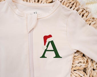 May include: A cream-colored long-sleeve onesie featuring a central zipper closure and an embroidered dark green letter A on the chest. The letter is topped with a red and white Santa hat design, adding a festive detail to the garment.