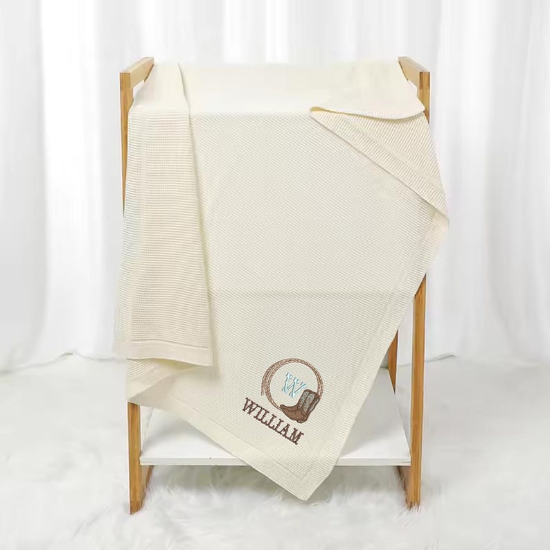 Puede incluir: Manta para beb&eacute; de color crema con textura de punto de gofre, exhibida en un estante de madera. La manta presenta un dise&ntilde;o bordado de una bota de vaquero y el nombre "WILLIAM" en hilo marr&oacute;n y azul.