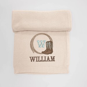 Puede incluir: Una manta de beb&eacute; de punto beige con tem&aacute;tica de vaquero. La manta presenta un dise&ntilde;o bordado de un lazo, una bota de vaquero y el nombre "WILLIAM" en letras marrones. Una letra "W" tambi&eacute;n est&aacute; bordada en el centro del lazo.