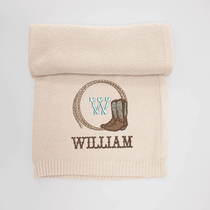 Puede incluir: Una manta de beb&eacute; de punto beige con tem&aacute;tica de vaquero. La manta presenta un dise&ntilde;o bordado de un lazo, una bota de vaquero y el nombre "WILLIAM" en letras marrones. Una letra "W" tambi&eacute;n est&aacute; bordada en el centro del lazo.