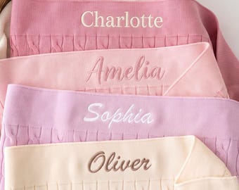 May include: Four folded knit blankets in shades of pink, light pink, lavender, and cream. Each blanket features a cable knit texture and is embroidered with a name in a different font and color: Charlotte, Amelia, Sophia, and Oliver.