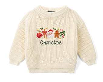 May include: A cream-colored, long-sleeved, ribbed knit sweater featuring an embroidered Christmas design. The graphic includes a red ornament, a bow, Santa Claus, a gingerbread man, and a candy cane, with the name Charlotte embroidered below.