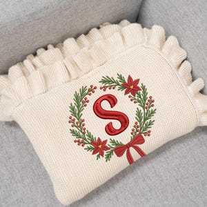 May include: Cream-colored knitted pillow with a decorative ruffle edge. The pillow features a red embroidered letter "S" inside a Christmas wreath with red berries, green leaves, and red poinsettias. A red bow is at the bottom.