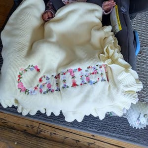 May include: Cream-colored baby blanket with the name "Carrie" embroidered in colorful floral letters. The blanket has a ruffled edge and a textured knit pattern. The blanket is draped over a baby in a car seat.