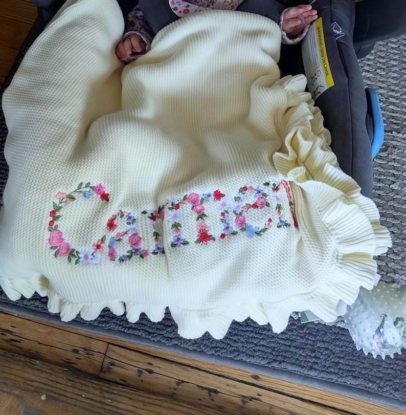 May include: Cream-colored baby blanket with the name "Carrie" embroidered in colorful floral letters. The blanket has a ruffled edge and a textured knit pattern. The blanket is draped over a baby in a car seat.