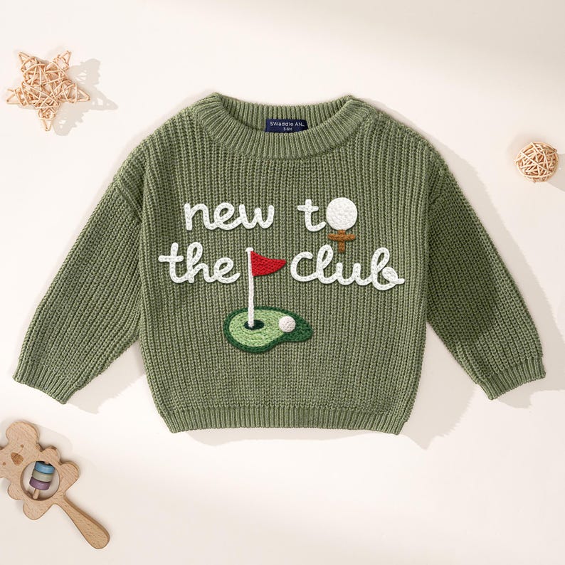 K&ouml;nnte beinhalten: Olivgr&uuml;ner Strickpullover mit dem wei&szlig;en Text "new to the club". Der Pullover hat einen gestickten Golfball, eine Flagge und Gr&uuml;n. Der Pullover ist ein Rundhalsausschnitt.