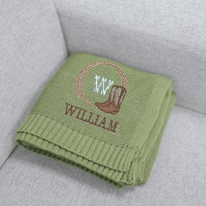 Puede incluir: Una manta de punto verde salvia con un dise&ntilde;o bordado marr&oacute;n. El dise&ntilde;o incluye un lazo, una bota de vaquero, la letra "W" y el nombre "WILLIAM". La manta est&aacute; doblada y apoyada sobre un sof&aacute; gris.