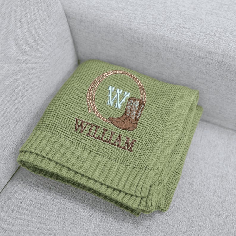 Puede incluir: Una manta de punto verde salvia con un dise&ntilde;o bordado marr&oacute;n. El dise&ntilde;o incluye un lazo, una bota de vaquero, la letra "W" y el nombre "WILLIAM". La manta est&aacute; doblada y apoyada sobre un sof&aacute; gris.