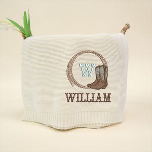 Puede incluir: Manta de punto color crema con un dise&ntilde;o bordado que presenta una bota de vaquero, la letra "W" y el nombre "WILLIAM". El dise&ntilde;o est&aacute; enmarcado por un c&iacute;rculo de cuerda. La manta tiene un borde acanalado.