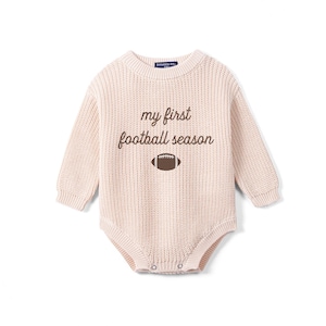 May include: A long-sleeved, light beige knit romper featuring a crew neckline and ribbed cuffs. The chest displays the brown embroidered text "my first football season" positioned above a small, brown and white graphic of a football.