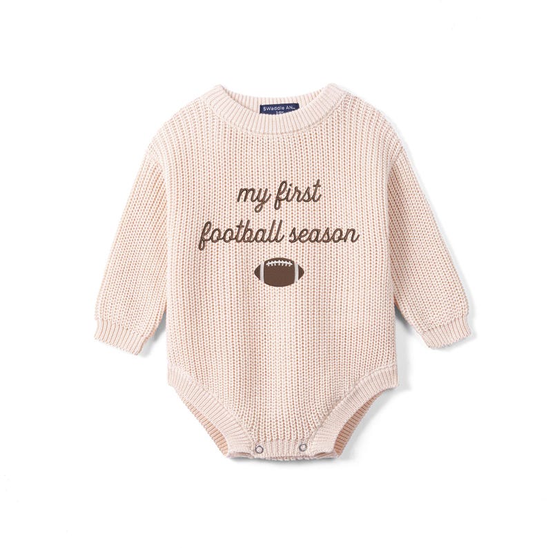 May include: A long-sleeved, light beige knit romper featuring a crew neckline and ribbed cuffs. The chest displays the brown embroidered text "my first football season" positioned above a small, brown and white graphic of a football.