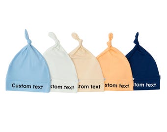 Könnte beinhalten: Fünf weiche, geknotete Baby-Mützen in Pastellfarben: Hellblau, Weiß, Beige, Pfirsich und Marineblau. Auf jeder Mütze steht "Custom text" auf der Vorderseite. Diese Mützen sind für Neugeborene konzipiert.