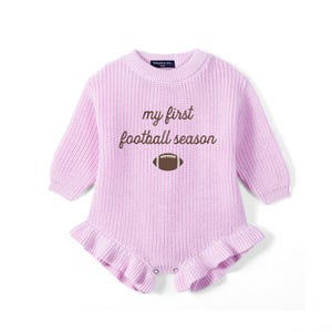 May include: A light purple long-sleeve knit romper featuring a ribbed crew neckline and cuffs. The chest displays the brown text 'my first football season' above a brown football graphic. The bottom hem is finished with a ruffled knit trim.