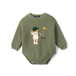 Peut inclure: Une barboteuse en tricot vert sauge à manches longues avec un col rond et une texture côtelée. Le centre présente un motif brodé représentant une silhouette portant une casquette verte et une tenue blanche, tenant un club de golf près d'un drapeau jaune.