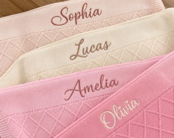 May include: Four stacked knitted blankets in shades of light pink, cream, and bright pink. Each blanket features a diamond-patterned texture and is embroidered with a name in cursive script: Sophia, Lucas, Amelia, and Olivia, respectively.