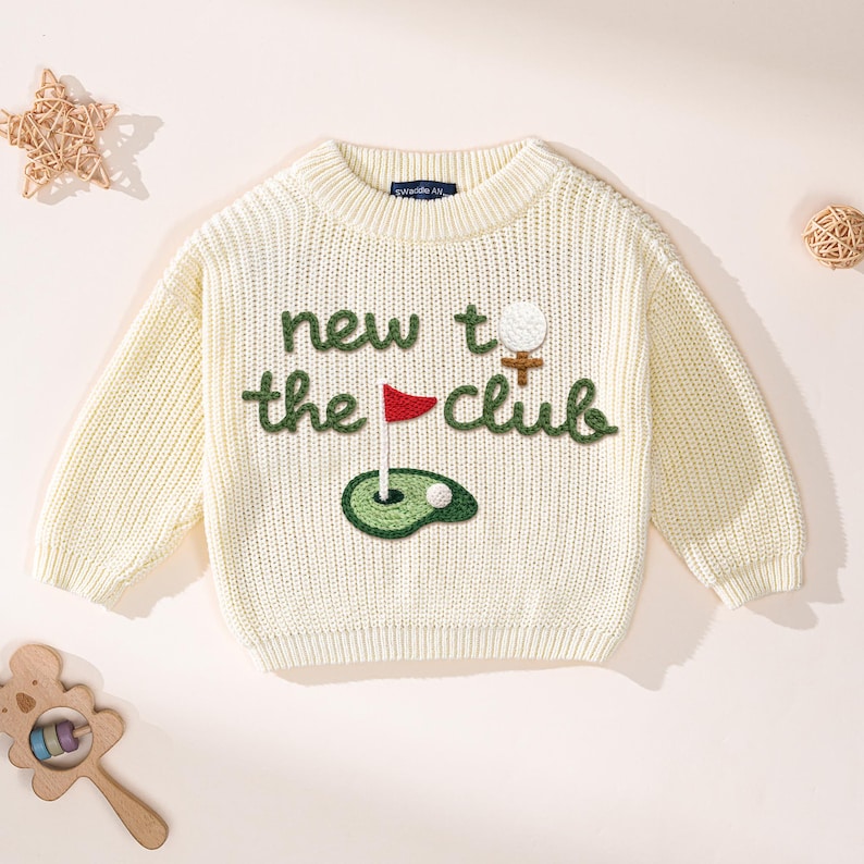 K&ouml;nnte beinhalten: Cremefarbener Strickpullover mit dem Text "new to the club" in Gr&uuml;n. Das Design umfasst einen Golfball, eine Fahne und ein Gr&uuml;n. Der Pullover hat lange &Auml;rmel und einen gerippten Rundhalsausschnitt. Ein verspieltes und bequemes Kleidungsst&uuml;ck f&uuml;r Kinder.