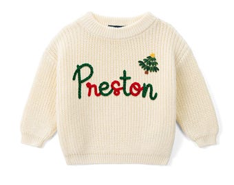 May include: A cream-colored, chunky knit sweater featuring the embroidered name Preston in alternating green and red script lettering. A small, embroidered green Christmas tree with a yellow star sits above the letter n.