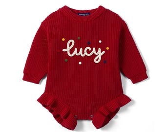 May include: A long-sleeved, deep red knit romper featuring a ribbed texture, a ruffled hem, and a crew neckline. The front displays the embroidered white script text "lucy" surrounded by small, colorful, scattered circular felt dots.