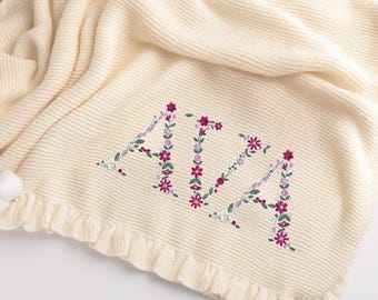 May include: Cream-colored knitted baby blanket with the name "AIA" embroidered in floral letters. The letters are adorned with purple and pink flowers and green leaves. The blanket has a ruffled edge and a pom-pom detail.