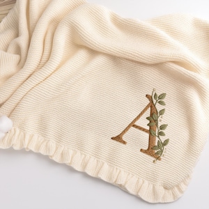May include: Cream-colored knitted baby blanket with a ruffled edge. The blanket features an embroidered letter "A" in brown with green leaf accents. The blanket is folded, showing its texture and design.