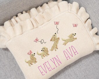 May include: Cream-colored knitted baby blanket with a ruffled edge. The blanket features embroidered tan-colored dachshunds and pink butterflies. The name "EVELYN AVA" is embroidered in pink script across the bottom.