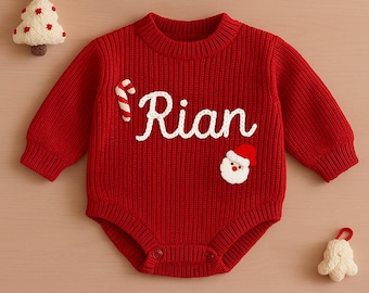 Pode incluir: Macacão de bebê de malha vermelha com o nome "Rian" em escrita branca. O macacão apresenta um bastão de doces e uma aplicação de rosto de Papai Noel. Uma pequena árvore de Natal e um sino também são visíveis.