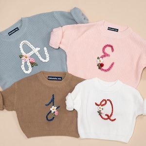 May include: Four pastel-colored knit sweaters with embroidered letters and floral accents. The sweaters are in blue, pink, brown, and white. Each sweater has a different letter: A, E, J, and Q. The brand name "Swaddle AN" is on the label.