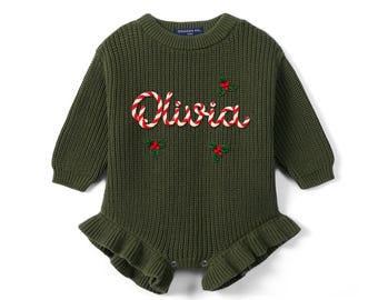 May include: An olive green long-sleeve knit romper featuring a crew neckline and a ruffled hem. The front displays the name Olivia in a red and white candy cane pattern, accented with embroidered green holly leaves and red berries.