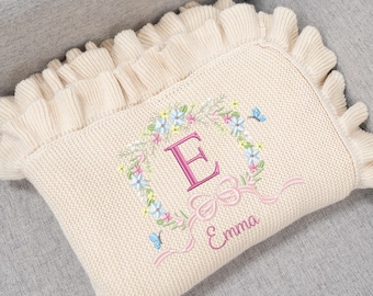 May include: Cream-coloured knitted baby blanket with a ruffled edge. The blanket features an embroidered floral wreath with a pink letter "E", a bow, and the name "Emma". Two small blue butterflies are also embroidered on the blanket.