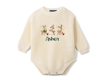May include: A cream-colored long-sleeve knit romper featuring a ribbed texture and a crew neckline. The chest displays an embroidered graphic of three reindeer with festive accents and the name Ashen in dark green script lettering.
