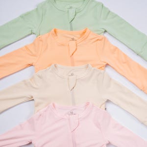 May include: Four pastel-colored baby sleepsuits are arranged diagonally. The sleepsuits are in shades of green, peach, cream, and pink. Each sleepsuit has a zipper down the front and long sleeves. The sleepsuits are made of a soft, comfortable material.