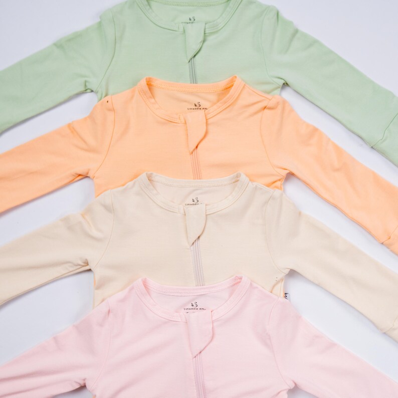 May include: Four pastel-colored baby sleepsuits are arranged diagonally. The sleepsuits are in shades of green, peach, cream, and pink. Each sleepsuit has a zipper down the front and long sleeves. The sleepsuits are made of a soft, comfortable material.