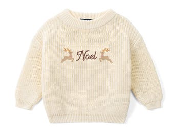 May include: A cream-colored, long-sleeved, chunky knit sweater featuring a crew neckline. The center of the chest displays an embroidered design with the word "Noel" in brown script, flanked by two small, light brown reindeer figures.