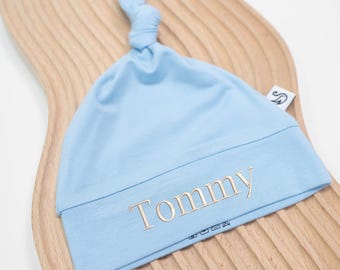 Könnte beinhalten: Hellblaues Baby-Mützchen mit geknotetem Oberteil. Der Name "Tommy" ist in Goldfaden auf dem umgeschlagenen Rand gestickt. Die Mütze ist aus weichem Material und für Säuglinge konzipiert.