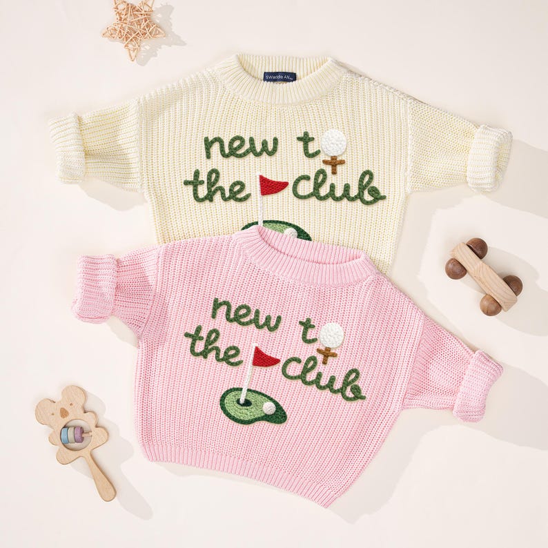 K&ouml;nnte beinhalten: Zwei Kinderpullover, einer cremefarben und einer rosa, mit dem gestickten Text "new to the club" und Golfmotiven. Ein Spielzeugauto und eine Rassel aus Holz sind ebenfalls im Bild.