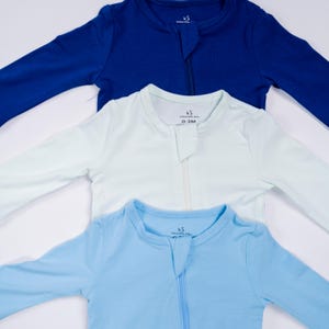 May include: Three baby sleepsuits in shades of blue and white are arranged in a layered composition. Each sleepsuit has long sleeves, a front zipper, and the text "Swaddle AR" on the collar. The sizes are 0-3M.