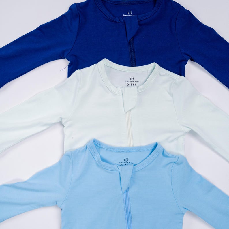 May include: Three baby sleepsuits in shades of blue and white are arranged in a layered composition. Each sleepsuit has long sleeves, a front zipper, and the text "Swaddle AR" on the collar. The sizes are 0-3M.