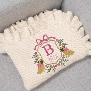 May include: Cream-colored knitted pillow with a ruffled edge. The pillow features an embroidered design with a pink bow, the letter "B", the name "BELLA", and two pug dog illustrations. The design is framed by a floral wreath.