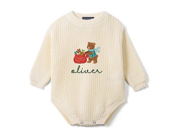 May include: A cream-colored long-sleeve knit romper featuring a ribbed texture and a crew neckline. The center displays an embroidered brown bear wearing a blue scarf, holding a red sack of gifts, with the name oliver written in green.