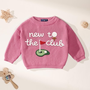K&ouml;nnte beinhalten: Ein rosa Strickpullover mit dem Schriftzug "new to the club" in wei&szlig;er Schrift. Der Pullover hat ein Golf-Motiv mit Golfball, Fahne und Gr&uuml;n. Der Pullover ist f&uuml;r ein Baby oder Kleinkind konzipiert.