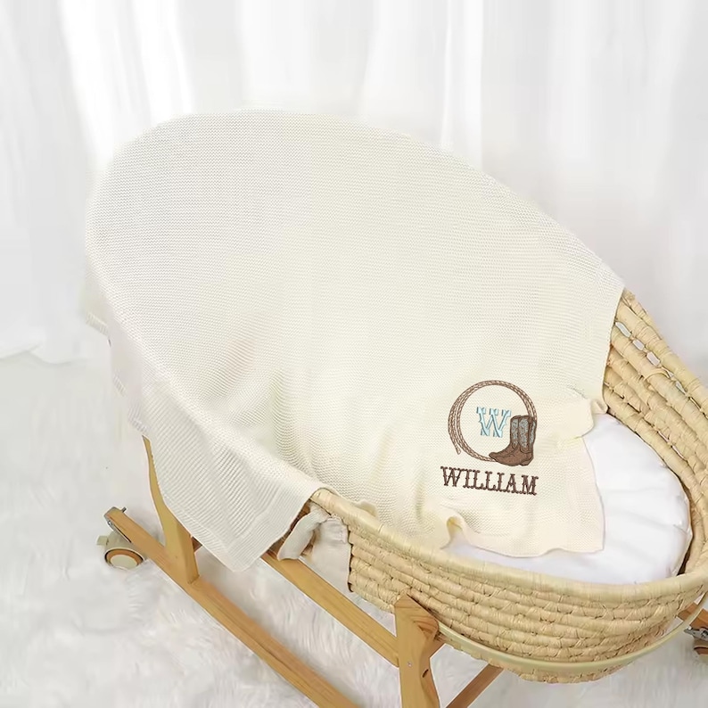 Puede incluir: Una manta de beb&eacute; color crema con un dise&ntilde;o de bota de vaquero y cuerda, personalizada con el nombre "WILLIAM" en marr&oacute;n. La manta est&aacute; colocada sobre una cuna de mimbre con un marco de madera.