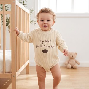 May include: A cream-colored long-sleeve knit baby bodysuit featuring the text "my first football season" in brown lettering, accompanied by a small brown football graphic centered on the chest. The garment has a ribbed texture and crew neckline.